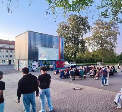 Im „Aktionssommer Zeitz“ finden Veranstaltungen wie Workshops, Kino, Konzerte und Sportevents am „Bluebox“-Gebäude statt, Foto: Stefan Scheel Jüngere und ältere Menschen schauen auf eine Kinoleinwand auf einem Platz in Zeitz.