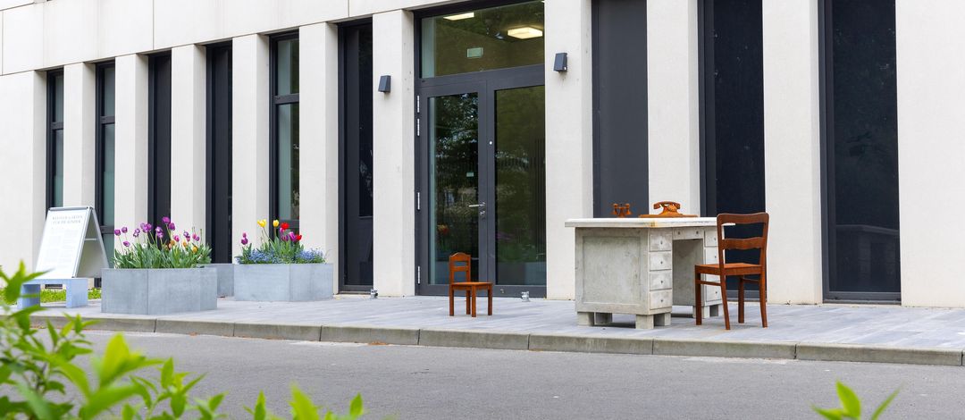 Ansicht des Mahnmal in Gedenken der "Kindereuthanasie" in Leipzig. Ein in Beton gegossener Schreibtisch mit Stuhl. Dem gegenüber steht ein Kinderstuhl. Im Hintergrund stehen drei Pflanzenkübel mit Tulpen.
