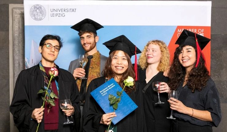 Five students pose after the graduation ceremony with a glass of wine, flowers and their diplomas.