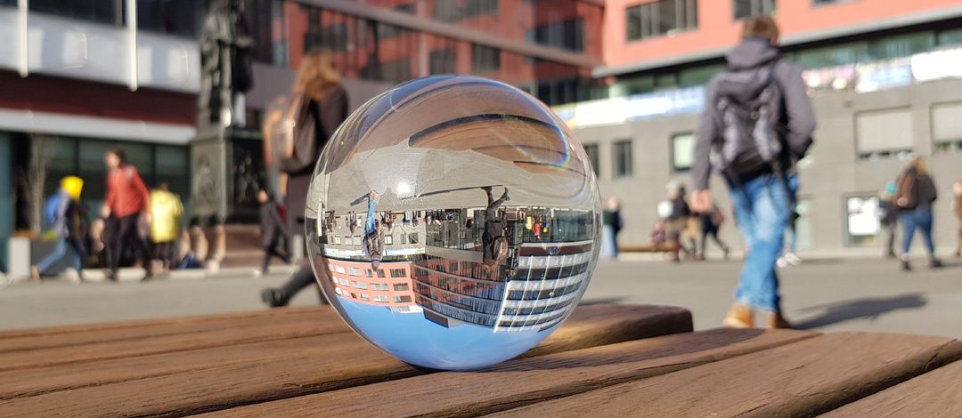 Farbfoto: Eine Glaskugel liegt auf einer Bank und in Ihr spiegeln sich Menschen und Gebäude aus dem Hintergrund