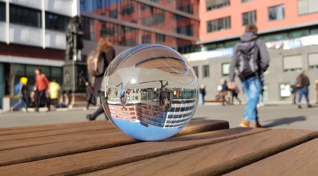 Farbfoto: Eine Glaskugel liegt auf einer Bank und in Ihr spiegeln sich Menschen und Gebäude aus dem Hintergrund