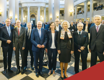 Farbfoto: 9 Menschen in festlicher Kleidung stehen in einer Reihe und lächeln in die Kamera. Im Hintergrund ist der Innenraum einer festlich beleuchten Kirche mit vielen Besuchern zu sehen.