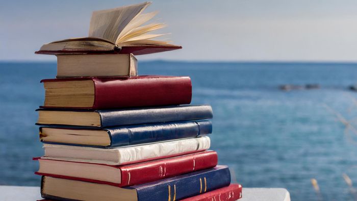 Ein Stapel Bücher in den Farben dunkelrot, weiß und dunkelblau liegt auf einem tisch, im Hintergrund das Meer.