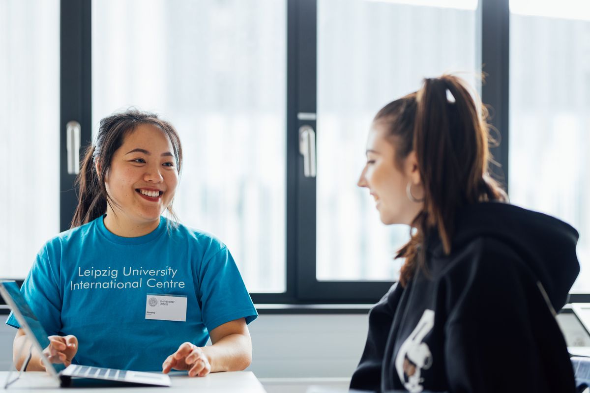 enlarge the image: Colour photo: A person at Leipzig University smiles at another person and shows them information on a laptop