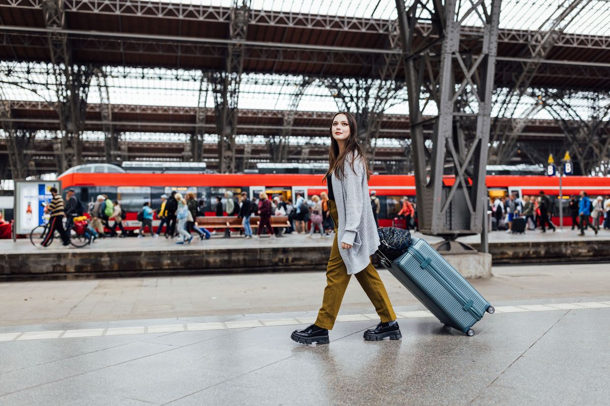 zur Vergrößerungsansicht des Bildes: Colour photo: A person walks with a suitcase along the station track. There is a train and many travellers in the background