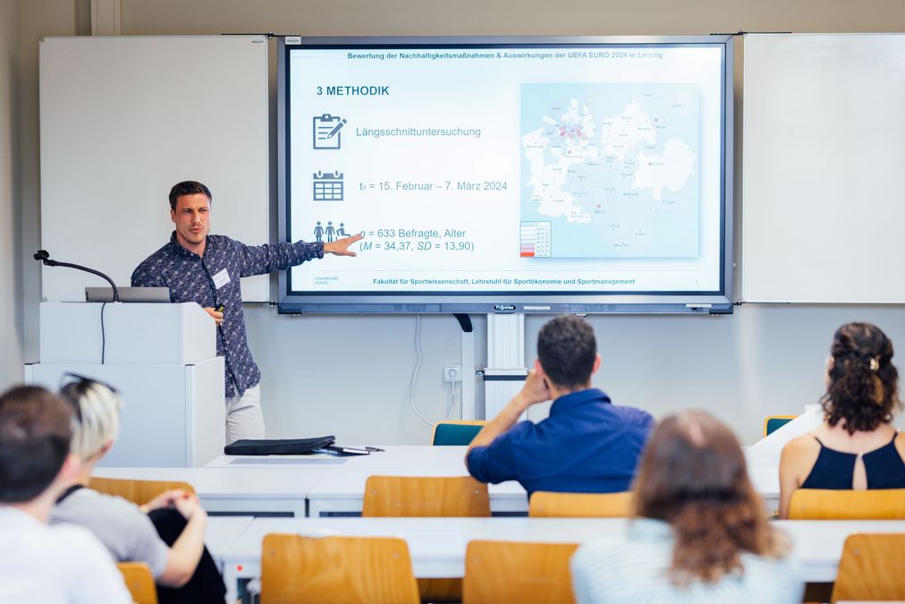 zur Vergrößerungsansicht des Bildes: Personen sitzen in einem Seminarraum, hinten im Bild ist eine digitale Tafel mit Folien zum Thema Nachhaltigkeit EUFA Euro 2024 in Leipzig