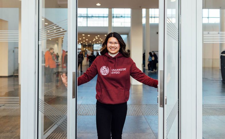 Farbfoto: Eine Frau mit roten Kaputzenpulli, auf dem groß das Logo der Universität Leipzig steht, öffnet freundlich lächelnd eine große Glastür.