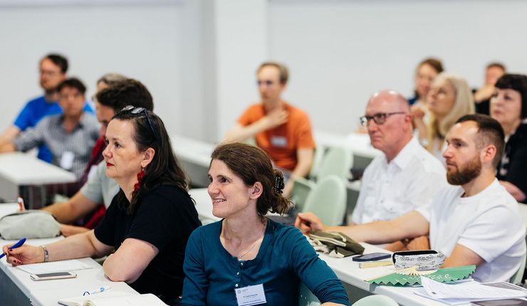 Auf vier Stuhlreihen verteilt in einem Seminarraum sitzen Menschen, die nach vorn blicken und einem Vortrag lauschen. Auf den Bänken sieht man Notizblöcke und Stifte. 