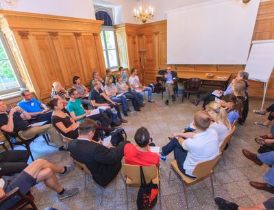 Die Fotografie zeigt eine Gruppe von ca. 30 Personen, die in einem Stuhlkreis zusammensitzen. Eine Person spricht und die anderen hören zu und machen sich Notizen.