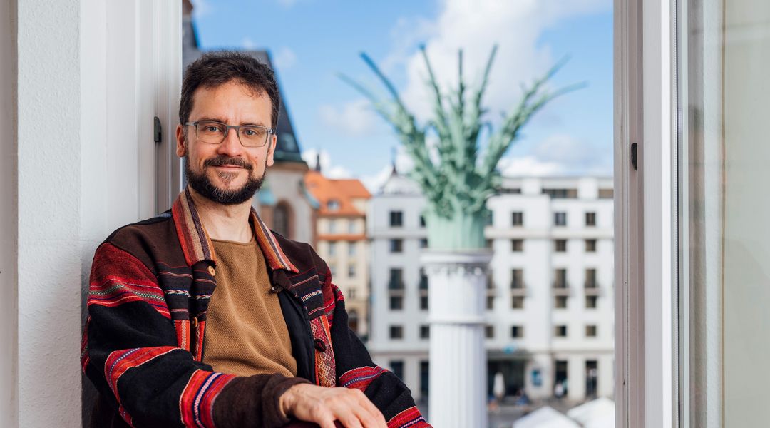 Zu sehen ist Dominik Becher im Fenster seines Büros, im Hintergrund ist der Nikolaikirchhof zu erkennen.