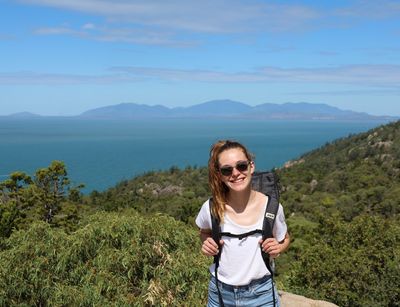 Studentin Estelle in Australien im Hintergrund Meer und Berge, sie steht inmitten von Grün 