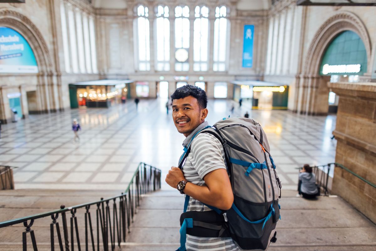 zur Vergrößerungsansicht des Bildes: Farbfoto: Eine Person mit Rucksack lächelt in die Kamera. Im Hintergrund befindet sich die Eingangshalle des Leipziger Bahnhofs.