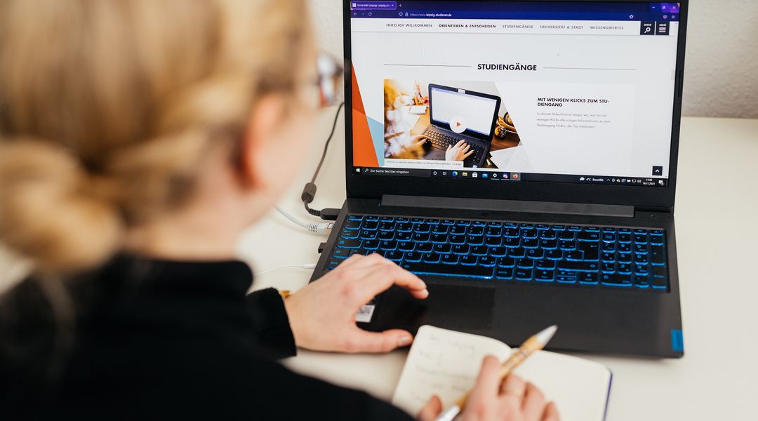 Farbfoto: Eine junge Frau sitzt mit dem Rücken zur Kamer vor einem Laptop und recherchiert auf der Website der Universität Leipzig nach passenden Studiengängen.