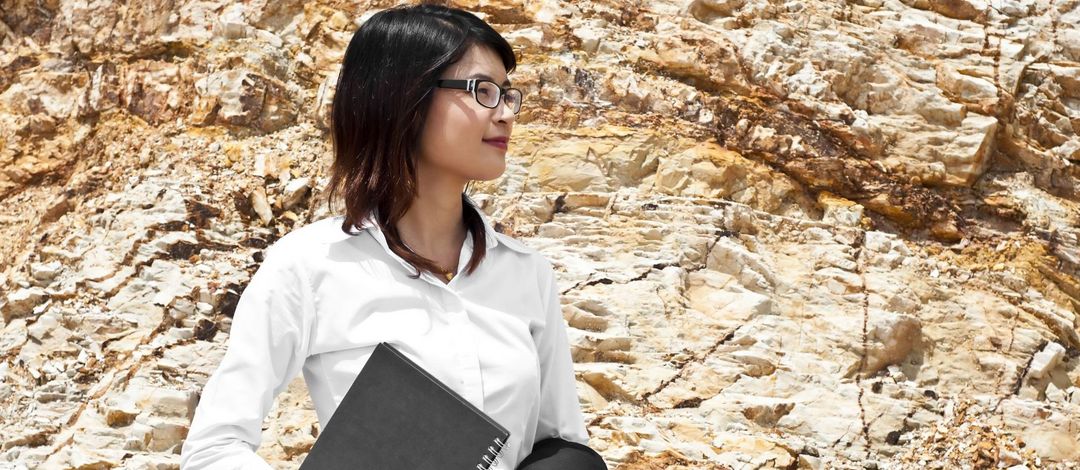 Farbfoto: Eine Frau mit weißer Bluse und Dokumentenmappe in der Hand stehtt vor einem braunen Felsen und lächelt. 