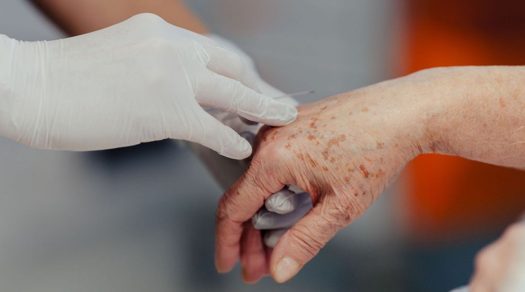 Farbfoto: Nahaufnahme von drei Händen. Zwei Hände mit weißen Gummihandschuhen halten eine andere Hand und versuchen eine Injektionsnadel einzuführen.. 