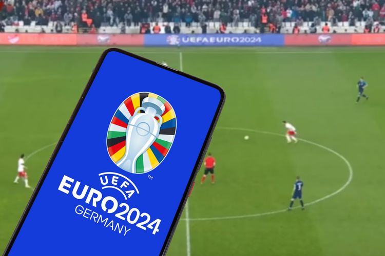 Zu sehen ist das Logo der Fußball-EM 2024 auf einem Smartphone. Im Hintergrund ist ein Spielfeld in einem Fußballstadion zu sehen.