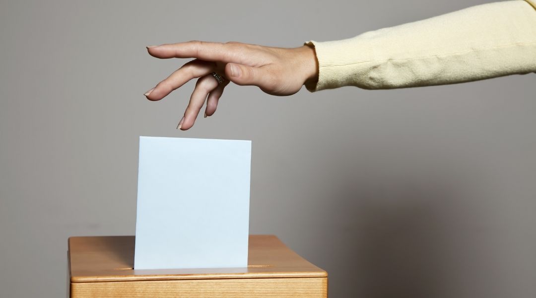 Farbfoto: Eine Hand wirft einen Stimmzettel in eine Wahlurne
